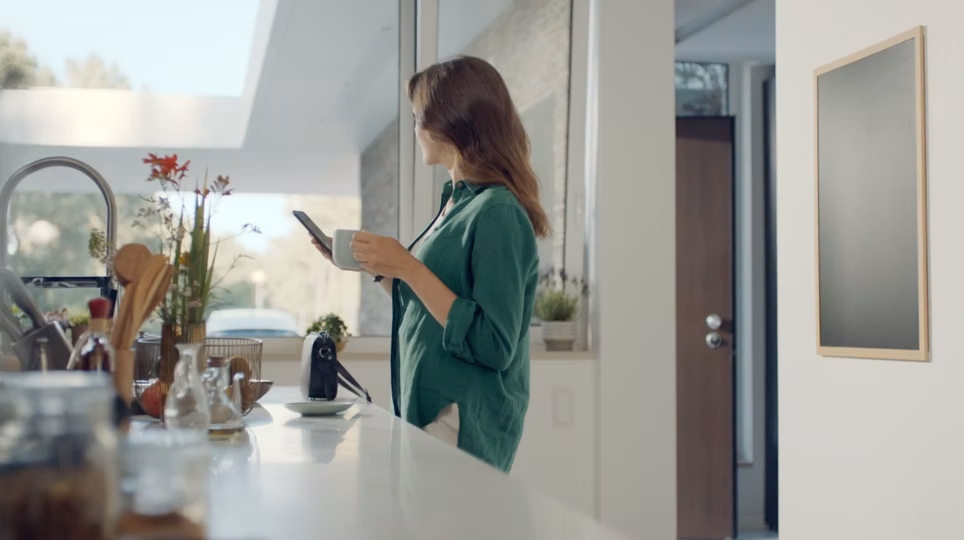 A woman at a kitchen counter, mug in one hand, smartphone in the other, a Hyundai parked in her driveway.   