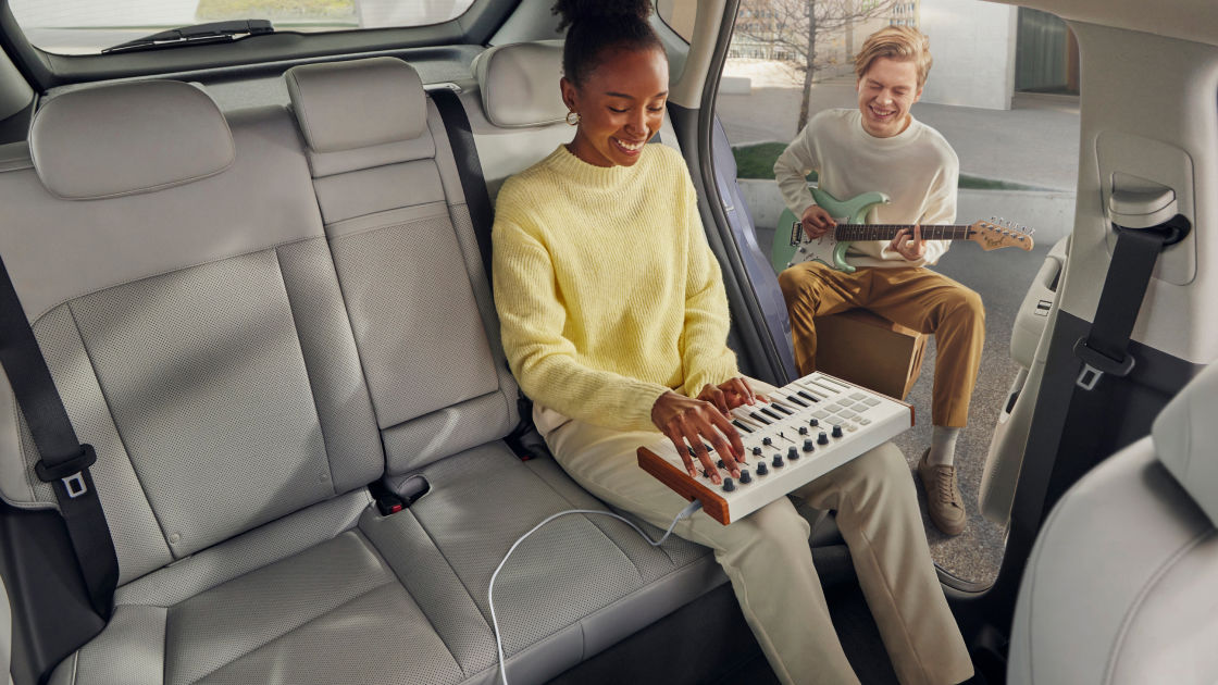 In the back seat of The all-new KONA with the door open, a woman is smiling and playing the electronic keyboard, while the man sitting outside The all-new KONA is smiling and playing the electronic guitar.